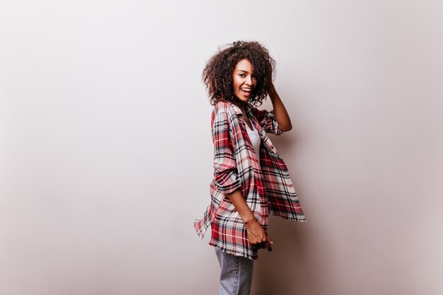 Chica adorable en ropa casual posando juguetonamente en blanco. Increíble mujer africana bien formada en camisa roja sonriendo