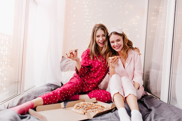 Chica adorable en traje de noche rojo de moda disfrutando de una pizza de queso con su hermana. Magníficas señoras sonrientes que pasan la mañana en la cama con comida rápida.