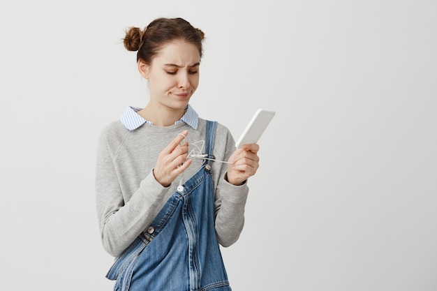 Foto gratuita chica adulta guapa que se irrita mientras intenta desenredar el cable de los auriculares. mujer en casual celebración smartphone con desentrañar auriculares estar molesto. reacciones humanas