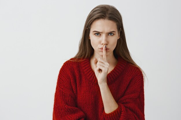 Chica advierte que no debemos contarle a nadie su secreto. Toma interior de preocupada seria mujer atractiva en suéter rojo suelto, callar, mostrando shh con el dedo índice sobre la boca, de pie sobre la pared gris