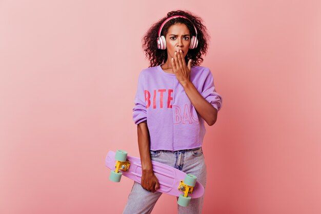 Chica africana bien vestida posando con longboard. Retrato de mujer negra elegante en auriculares de pie en rosa con expresión de cara de sorpresa.