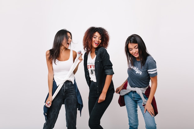 Chica africana despreocupada divirtiéndose con amigos de moda sonriendo. Emocionada modelo de mujer negra de pie con los ojos cerrados mientras damas morenas bailan al lado.