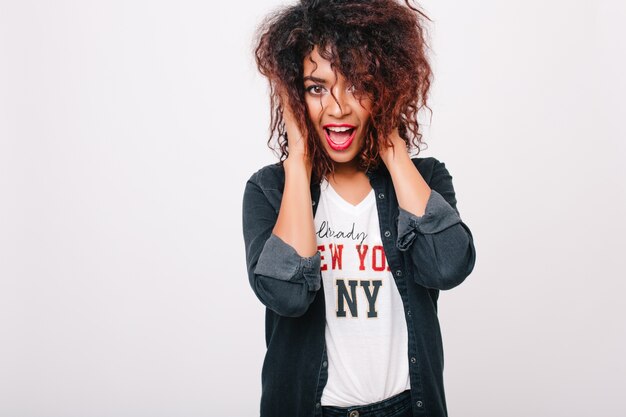 Chica africana extática con rizos de color marrón oscuro posando con la boca abierta y expresión de la cara feliz. Retrato de interior de dama mulata sorprendida pero dichosa con camisa blanca con estampado.