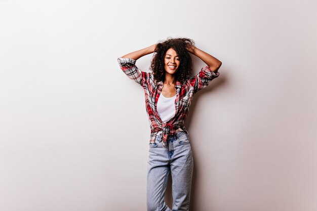 Chica africana increíble delgada expresando emociones positivas en blanco. elegante mujer riendo con pelo corto y rizado posando en camisa a cuadros.