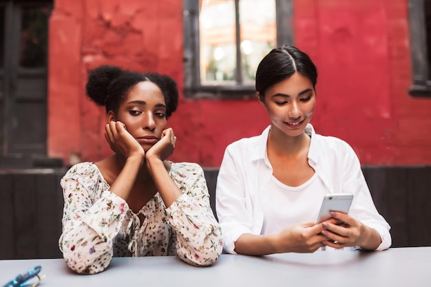 Foto gratuita chica africana molesta mirando tristemente a un lado mientras una chica asiática cerca felizmente usando el teléfono celular en el acogedor patio de la cafetería