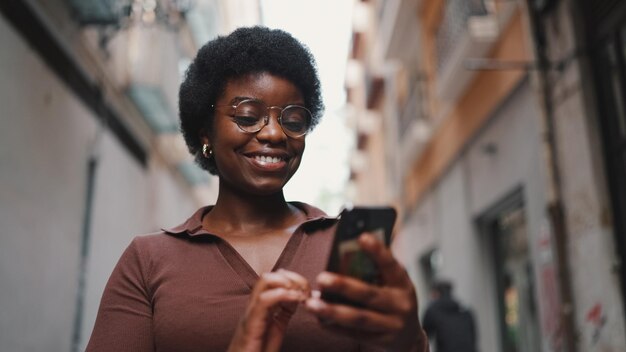 Chica afro joven con gafas que parece feliz enviando mensajes de texto con frien