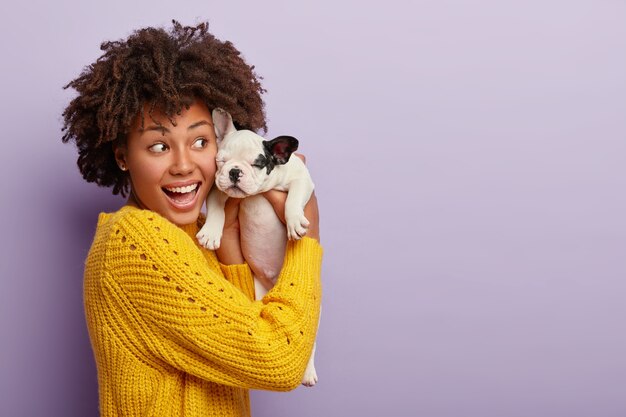 Chica afro positiva en jersey amarillo sostiene un pequeño cachorro con oreja negra, juega con una linda mascota, se siente llena de energía y complacida