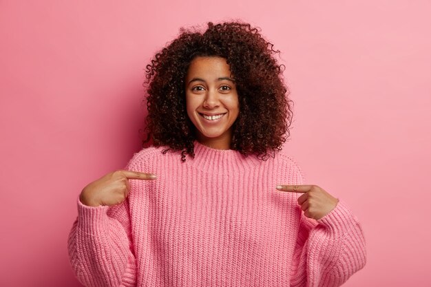 La chica afro positiva se señala a sí misma, indica el pecho, se ve jactanciosa, se viste con un jersey de punto de gran tamaño, comparte buenas noticias, sonríe agradablemente, aislada sobre una pared rosa