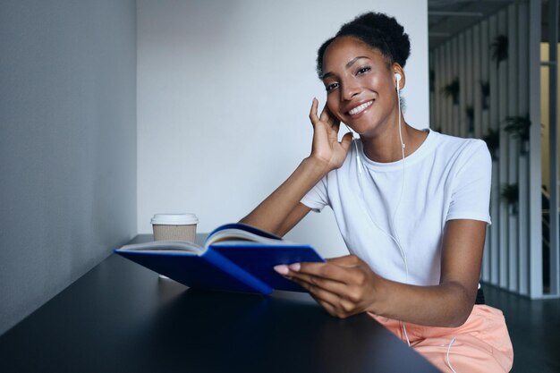 Chica afroamericana bastante informal con auriculares con libro y café mirando alegremente a la cámara en un espacio de trabajo conjunto moderno
