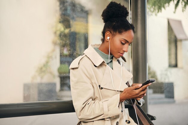 Una chica afroamericana bastante informal con una elegante gabardina y auriculares usando un teléfono celular en la parada de autobús