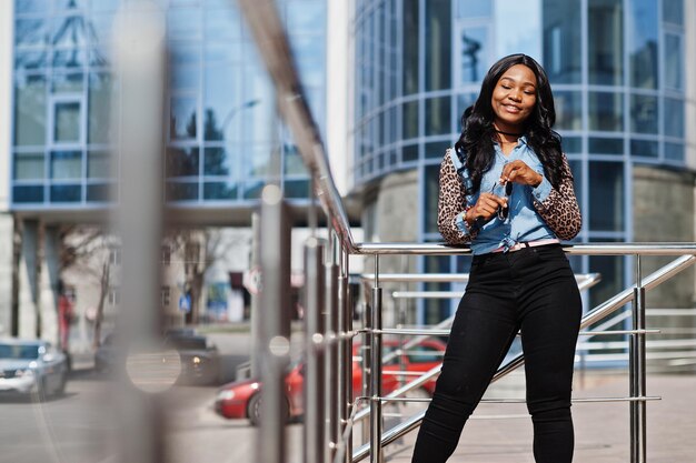 Chica afroamericana hipster con camisa de jeans con mangas de leopardo posando en la calle contra un edificio de oficinas moderno con ventanas azules