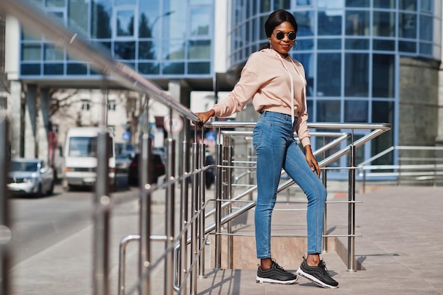 Chica afroamericana hipster con capucha rosa, gafas de sol y jeans posando en la calle contra el edificio de oficinas con ventanas azules