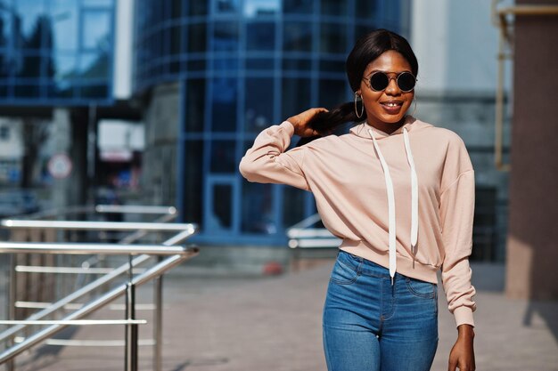 Chica afroamericana hipster con capucha rosa, gafas de sol y jeans posando en la calle contra el edificio de oficinas con ventanas azules