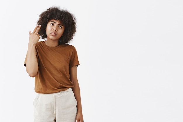 Chica afroamericana insatisfecha melancólica con peinado rizado en camiseta marrón muriendo de aburrimiento y molestia con los párpados rodantes haciendo disparos de pistola en la cabeza