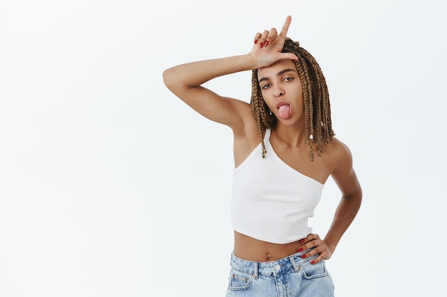 Chica afroamericana joven descarada burlándose de alguien, mostrando gesto de perdedor en la frente y la lengua