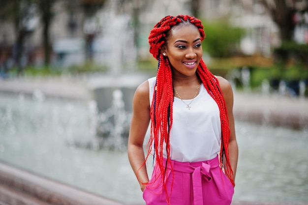 Chica afroamericana de moda en pantalones rosas y rastas rojas posadas al aire libre contra fuentes