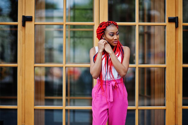 Chica afroamericana de moda en pantalones rosas y rastas rojas posadas al aire libre