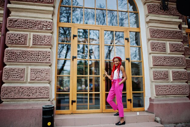 Chica afroamericana de moda en pantalones rosas y rastas rojas posadas al aire libre