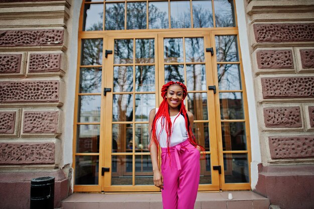 Chica afroamericana de moda en pantalones rosas y rastas rojas posadas al aire libre