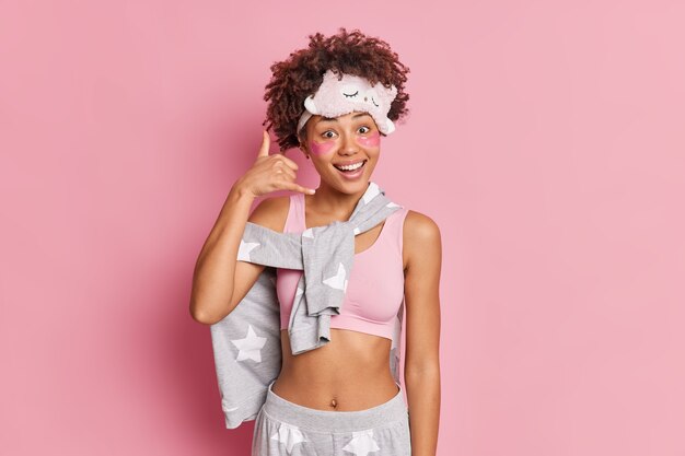 Chica afroamericana positiva hace que el gesto me devuelva la llamada sonríe felizmente vestida con ropa de dormir muestra un vientre plano que aplica parches de hidrogel debajo de los ojos aislados sobre una pared rosa