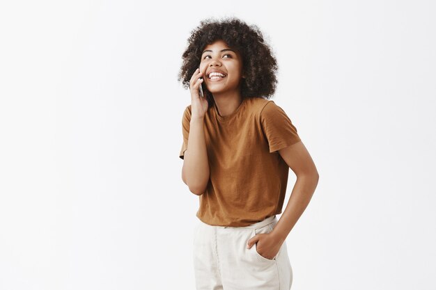 Chica afroamericana relajada divirtiéndose hablando casualmente a través de un teléfono inteligente mirando hacia arriba con carefre alegre sonrisa sosteniendo la mano en el bolsillo de pie