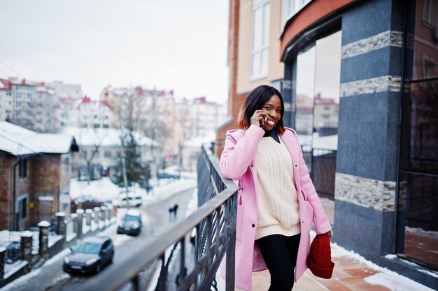 Chica afroamericana con sombrero rojo y abrigo rosa en la calle de la ciudad contra la construcción en el día de invierno con teléfono móvil