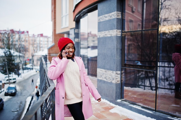 Chica afroamericana con sombrero rojo y abrigo rosa en la calle de la ciudad contra la construcción en el día de invierno con teléfono móvil