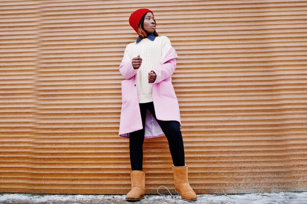 Chica afroamericana con sombrero rojo y abrigo rosa contra persianas naranjas