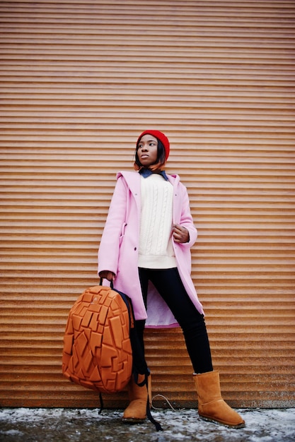 Chica afroamericana con sombrero rojo y abrigo rosa con mochila contra persianas naranjas