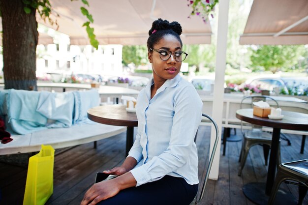 Chica afroamericana usa gafas con teléfono móvil sentado en un café al aire libre
