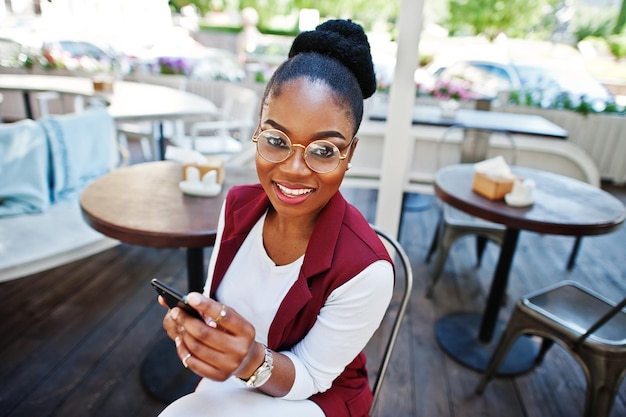 Chica afroamericana usa gafas con teléfono móvil sentado en un café al aire libre