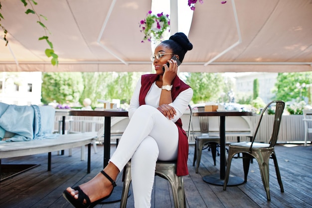 Chica afroamericana usa gafas con teléfono móvil sentado en un café al aire libre