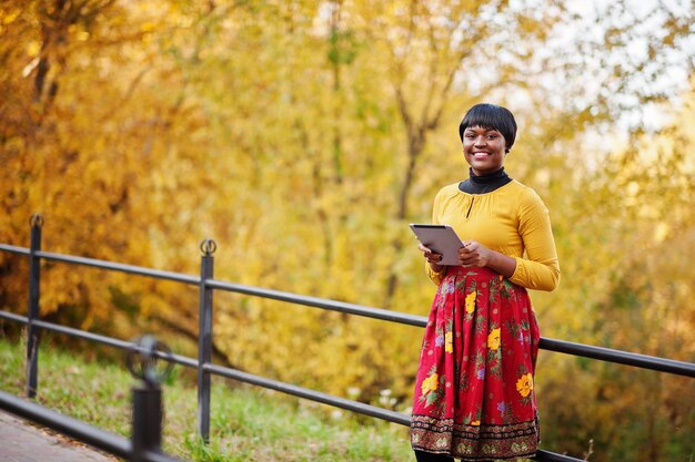 Chica afroamericana en vestido amarillo y rojo en el parque de otoño dorado con tableta en las manos