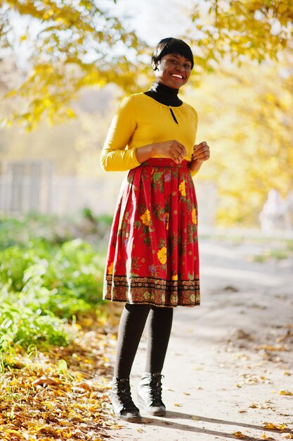 Chica afroamericana en vestido amarillo y rojo en el parque de otoño dorado