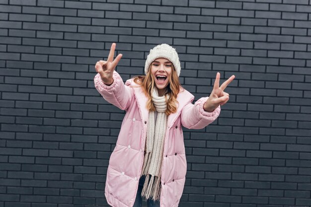 Chica agraciada en elegante chaqueta de invierno divirtiéndose al aire libre. Foto de magnífica mujer rubia con gorro de punto blanco.