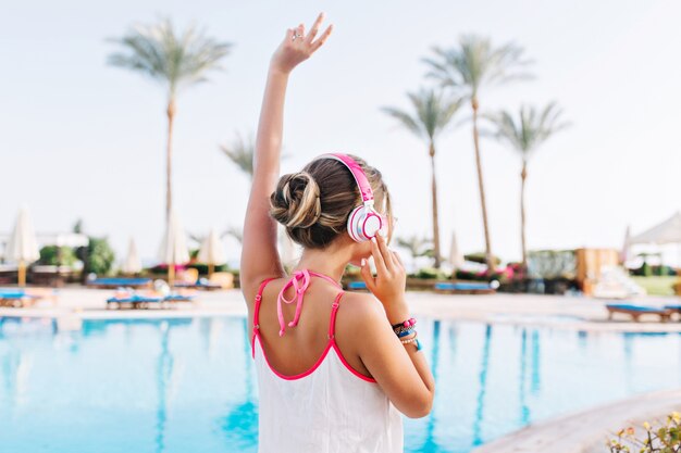 Chica agraciada con piel bronceada con camiseta blanca y posando con la mano cerca de la piscina
