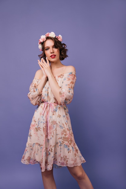 Chica agraciada en vestido de moda de pie. Mujer interesada con flores en el pelo ondulado posando.