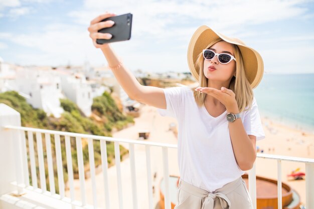 Chica agradable con gafas de sol y sombrero envía besos mientras hace selfie en el mar