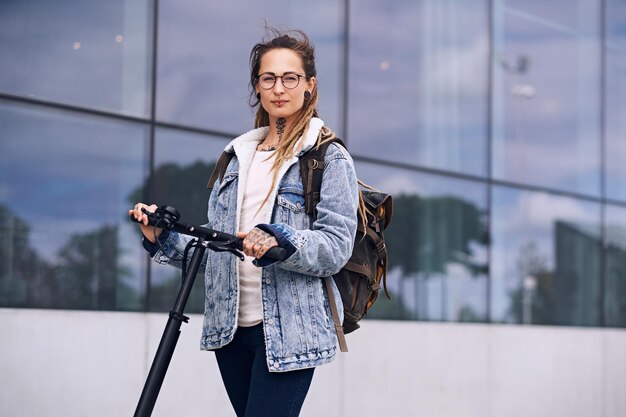 Una chica agradable y pensativa con gafas y tatuajes en el cuello y las manos conduce una moto eléctrica.