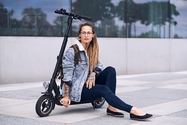 Una chica agradable y pensativa con gafas y tatuajes en el cuello y las manos se está relajando en su propia moto.