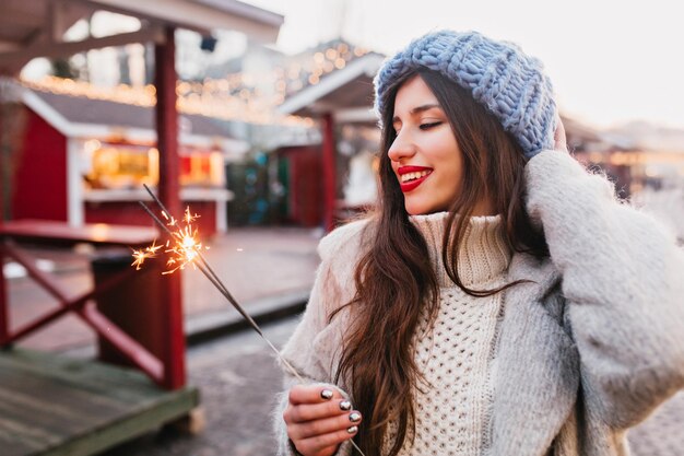 Chica alegre en abrigo gris mirando la luz de Bengala con una sonrisa en el día de invierno. Encantadora mujer caucásica con sombrero azul de pie con bengala en la calle, esperando la Navidad.