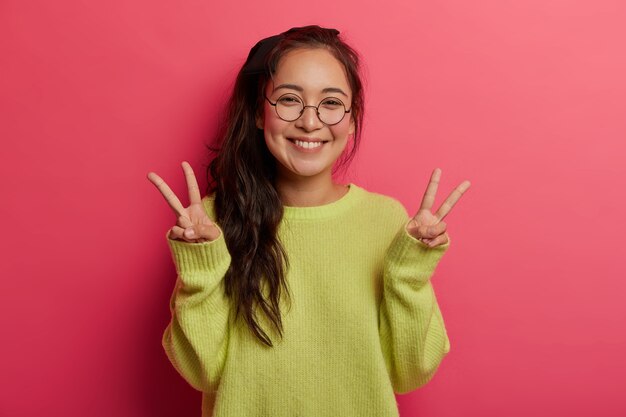 La chica alegre y afortunada muestra un gesto de victoria o paz, disfruta de un día increíble, envía vibraciones positivas, tiene una sonrisa con dientes, cabello oscuro peinado en una cola de caballo