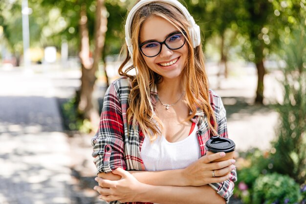 Chica alegre en auriculares blancos sonriendo a la naturaleza. Tiro al aire libre de increíble modelo femenino con taza de café escuchando música.