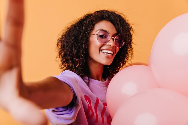 Chica alegre con cabello ondulado posando con entusiasmo en naranja. Jocund mujer africana haciendo selfie con globos de helio.