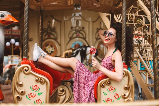 Chica alegre con cabello oscuro y rizado con gafas de sol y vestido sosteniendo caramelos en la mano y montando en carrusel mientras pasa felizmente el tiempo en el parque de diversiones