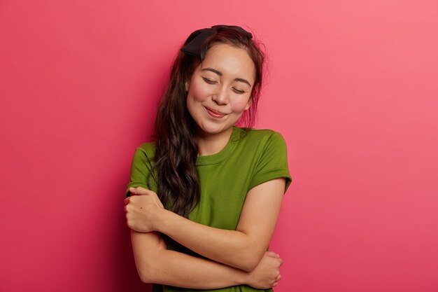 chica alegre con cabello oscuro tiene un estado de ánimo romántico, se abraza, inclina la cabeza, mantiene los ojos cerrados, le gusta su propio cuerpo, usa una camiseta verde informal, posa en interiores.