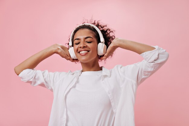 Chica alegre en camisa blanca y auriculares posando en la pared rosa