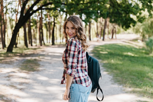 Chica alegre en camisa a cuadros y jeans de pie en el parque. Mujer inspirada con mochila de cuero sonriendo en primavera.