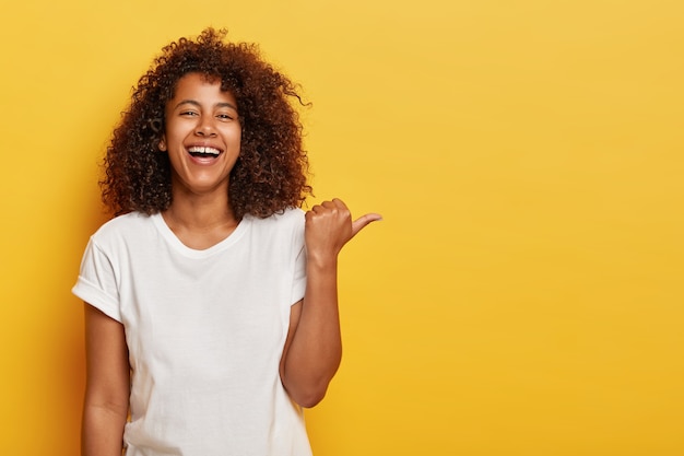 Una chica alegre y encantadora señala con el pulgar hacia el costado, se ríe alegremente, tiene una sonrisa brillante, demuestra algo genial, se siente divertida, está de buen humor, usa una camiseta blanca, posa contra la pared amarilla