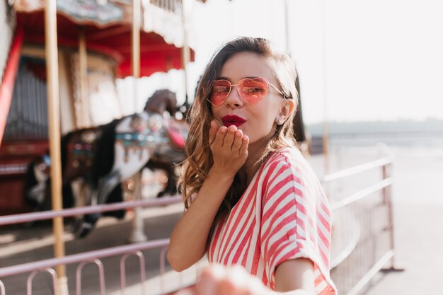 Chica alegre en gafas de sol de corazón pasar tiempo en el parque de atracciones. Mujer muy elegante enviando beso al aire mientras se relaja en verano.